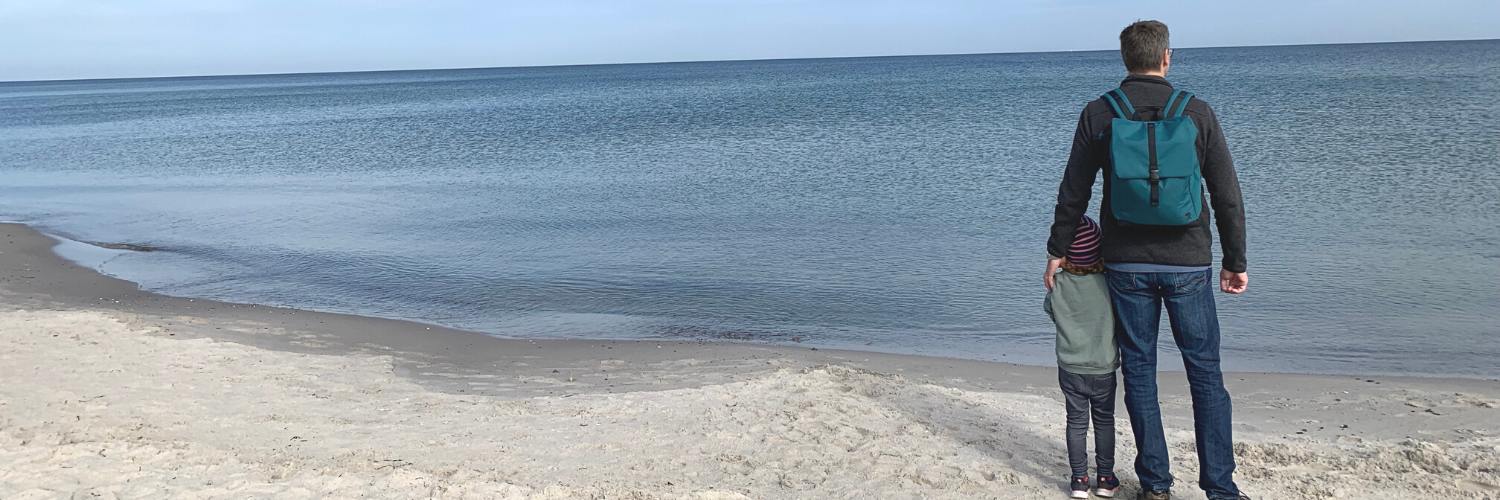 Vater mit dem FUCHS und REBELL Rucksack MATS in petrolblau steht mit seiner Tochter am Strand von Boltenhagen und blickt auf die Ostsee